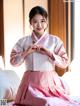 A woman in a pink and white hanbok sitting on a bed.